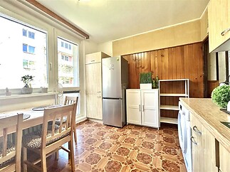 Světlá kuchyň s dřevěnými obklady, parketovou podlahou a velkými okny. Nábytek zahrnuje vestavěné skříně a moderní spotřebiče.