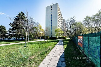 Vysoký panelový bytový dům s dobře udržovaným trávníkem a chodníkem, nacházející se v klidné rezidenční oblasti.