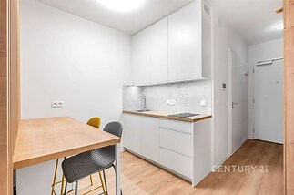 Kompaktní kuchyň s moderním nábytkem, bílými skříňkami a dřevěnou podlahou, doplněná světlým obkladem a jídelním koutem pro dva.