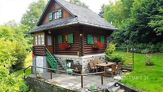 Dřevěná chalupa s kamenným základem, terasa s venkovním posezením, obklopená zelení, rustikální styl, klidná lokalita v přírodě.