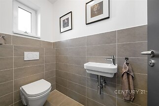 Moderní koupelna s dlažbou, podomítkovým WC a malým umyvadlem. Světlý prostor s oknem a minimalistickým designem.