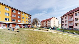 Dům s barevnou fasádou, parkoviště a dětské hřiště v klidném prostředí, vhodné pro rodiny. Okolí nabízí zelené plochy pro relaxaci.