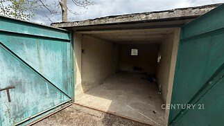 Opuštěná garáž s betonovou podlahou, starší dřevěná vrata, malá okna, potřebná rekonstrukce, vhodná pro skladování nebo parkování.