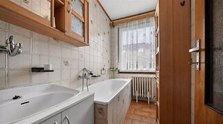 Koupelna s keramickými dlaždicemi, umyvadlem a vanou, doplněná dřevěnými skříňkami, světlo z velkého okna, vytápění radiátorem.