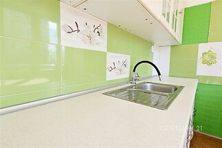 Moderní kuchyň s bílou pracovní deskou, zelenými dlaždicemi a nerezovým dřezem, světlé prostředí, stylová keramika.