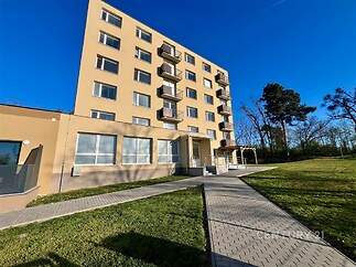 Moderní bytový dům s velkými okny, rekonstruovanou fasádou a prostornými balkony ve světlém odstínu, obklopený zelení a zpevněným chodníkem.