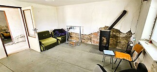 Průchozí obývací pokoj s krbovými kamny a betonovou podlahou, vyžadující rekonstrukci, dodává prostoru rustikální atmosféru.