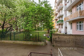 Udržovaná zahrada ve vnitrobloku s vzrostlými stromy, zeleným trávníkem a okolními bytovými domy, ideální pro relaxaci v přírodním prostředí.