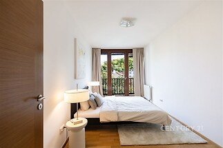 Světlá ložnice s parketovou podlahou, velká okna s výhledem do zahrady, moderní nábytek a čistý design vytvářejí útulný a elegantní prostor.