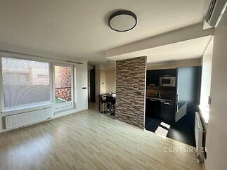 Světlý obývací pokoj s parketovou podlahou, velká okna a moderní kuchyň se zástěnou z kamenů, vytváří útulný a stylový prostor.