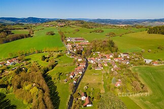 Letecký pohled na malebnou vesnici obklopenou zelenými loukami a kopci, ideální lokalita pro klidné bydlení v přírodě s krásnými výhledy.
