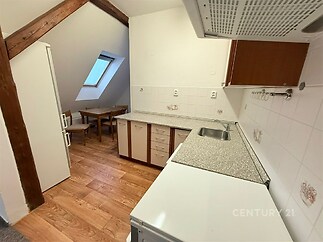 Malá kuchyň s dřevěnou podlahou, šikmým stropem a střešním oknem. Moderní vybavení a bílé obklady dodávají světlý vzhled.