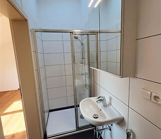 Malá koupelna s moderním sprchovým koutem, světlými dlaždicemi a velkým zrcadlem, poskytuje elegantní a funkční prostor v rekonstruovaném bytě