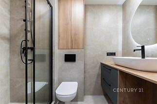 Moderní koupelna s dlažbou, elegantním umyvadlem a sprchovým koutem. Dřevěný obklad a černé doplňky vytvářejí stylový vzhled.