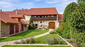 Upravená zahrada s dlážděnými chodníky, travnatou plochou a vzrostlými jehličnany. Tradiční dům s kamennými prvky a červenou střechou.