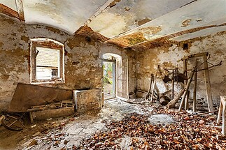 Zchátralá místnost s klenutým stropem a cihlovým omítaným povrchem, dveře vedou na dvůr, neudržovaný prostor s historickými prvky.