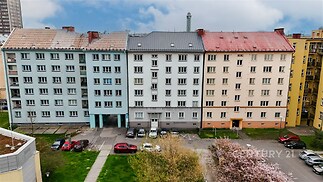 Pohled na fasády panelových domů s barevnými omítkami v městské zástavbě. Domy mají různé barvy a běžné stavební prvky.