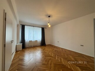 Prostorný obývací pokoj s parketovou podlahou, velká okna s závěsy, moderní osvětlení, světlé stěny a čistý minimalistický design.