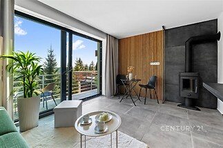 Světlý obývací pokoj s moderními krbovými kamny, velkými okny a dlažbou. Elegantní design nabízí výhled na balkón a zeleň.