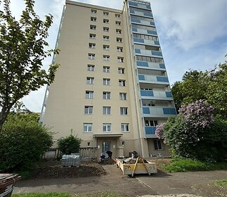 Vysoký bytový dům s modernizovanou fasádou, obklopený zelení a květinami, ideální pro rodinné bydlení v klidné lokalitě.
