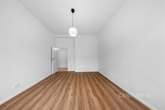 Prostorný pokoj s moderní dřevěnou podlahou, vysokým stropem a jednoduchým závěsným svítidlem, světlá výmalba a otevřené dveře do další místnosti.