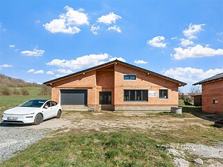 Moderní rodinný dům s parkovacím stáním, velkými okny a neomítanými cihlami. Nachází se v klidné lokalitě s přírodním pozadím.