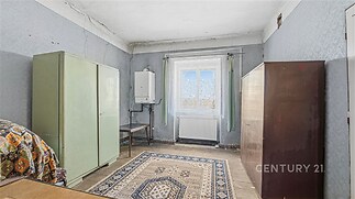 Ložnice s dlažbou, starším nábytkem a velkým oknem, které poskytuje přirozené světlo. Místnost vyžaduje rekonstrukci.