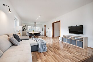 Světlý obývací pokoj s parketovou podlahou, moderní dekorací a velkými okny. Otevřený prostor propojený s kuchyní a jídelnou nabízí pohodlí.