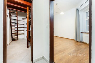 Průchod do světlého pokoje s parketovou podlahou a velkými okny vedle úložného prostoru s dřevěnými regály a schodištěm