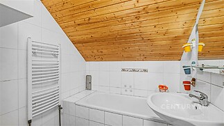 Koupelna s bílými dlaždicemi a dřevěným obložením stropu, obsahující vanu, umyvadlo a moderní topný žebřík, vytváří útulný dojem.