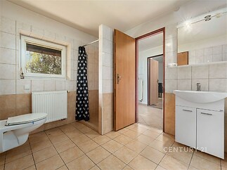 Prostorná koupelna s dlažbou, sprchovým koutem a umyvadlem, moderní design, přirozené světlo přichází velkým oknem, světlé dlaždice.