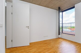 Světlý pokoj s parketovou podlahou a velkým oknem, které nabízí krásný výhled. Moderní interiér s jednoduchými liniemi a stropem z pohledového betonu.