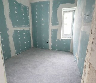 Rekonstruovaný pokoj s nedokončenými stěnami, velkým oknem a světlou podlahou, ideální pro přizpůsobení interiéru podle vlastních představ