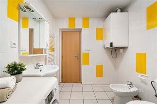 Moderní koupelna s dlažbou a žlutými akcenty, vybavená bidetem a pračkou. Světlé stěny a vestavěná skříňka vytvářejí prostorný dojem.