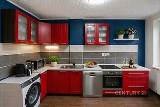 Moderní kuchyň s červenými skříňkami, nerezovými spotřebiči a tmavou parketovou podlahou. Světlá, s velkým oknem a stylovým designem.