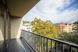 Prostorný balkon s dlažbou, nabízí výhled na městskou architekturu a zeleň. Moderní zábradlí a velká okna dotvářejí světlou atmosféru.