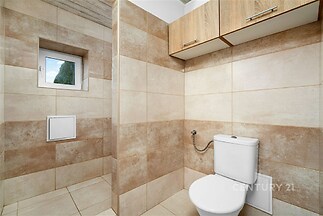 Moderní koupelna s dlažbou, světlými obklady a dřevěnými skříňkami. Kompaktní prostor s malým oknem poskytuje praktické uspořádání.