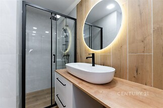 Moderní koupelna s dřevěným obložením, prosvětlené zrcadlo, skleněný sprchový kout a stylová černá baterie, elegantní design.