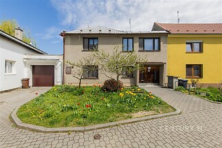 Rodinný dům s tradiční fasádou, upravenou zahradou a garáží. Přístupová cesta je z dlažby. Dům má moderní okna a udržovaný exteriér.