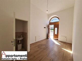 Světlý vstupní prostor s parketovou podlahou a vysokým stropem, vedoucí na terasu přes dřevěné dveře. Moderní interiér s přirozeným osvětlením.