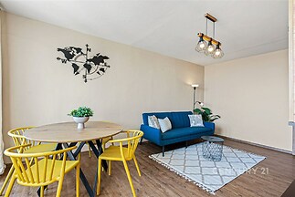 Světlý obývací pokoj s moderní výzdobou, dřevěnou podlahou a stylovým nábytkem. Velká okna umožňují dostatek přirozeného světla.