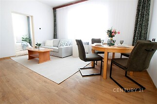 Světlý obývací pokoj s parketovou podlahou, velkými okny a moderním nábytkem. Komfortní prostor s jídelním stolem a pohovkou.