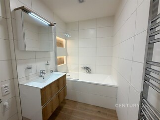 Moderní koupelna s bílou dlažbou, dřevěným umyvadlovým pultem a vanou. Světlý a čistý design s praktickým zrcadlem a topným žebříkem.