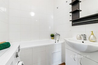 Kompaktní koupelna s bílými dlaždicemi, praktickou vanou a moderním umyvadlem, nabízí úložné prostory a minimalistický design.