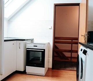 Kuchyň v podkroví, světlá dřevěná podlaha, střešní okno, moderní spotřebiče, bílá kuchyňská linka.