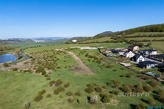 Výhled na stavební pozemek s možností výstavby, obklopený zelenými kopci a moderními rodinnými domy, klidná lokalita.
