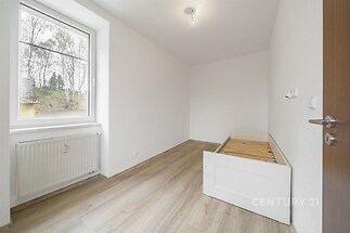 Světlý pokoj s dřevěnou podlahou, velkým oknem a moderním designem, ideální pro rekonstrukci nebo zařízení jako ložnice.