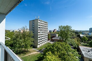 Pohled z balkonu s moderním zábradlím na sídliště, obklopené zelení a panelovými domy, ideální pro klidné bydlení ve městě.