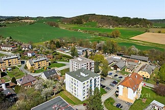 Pohled na obytnou čtvrť s moderními bytovými domy a zelenými zahradami, okolní příroda poskytuje klidnou atmosféru blízko městské oblasti.