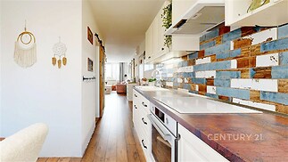 Moderní kuchyň s dřevěnou podlahou, barevný obklad stěn a vestavěné spotřebiče. Světlý interiér s dlouhou pracovní deskou a dekorativním prvky.